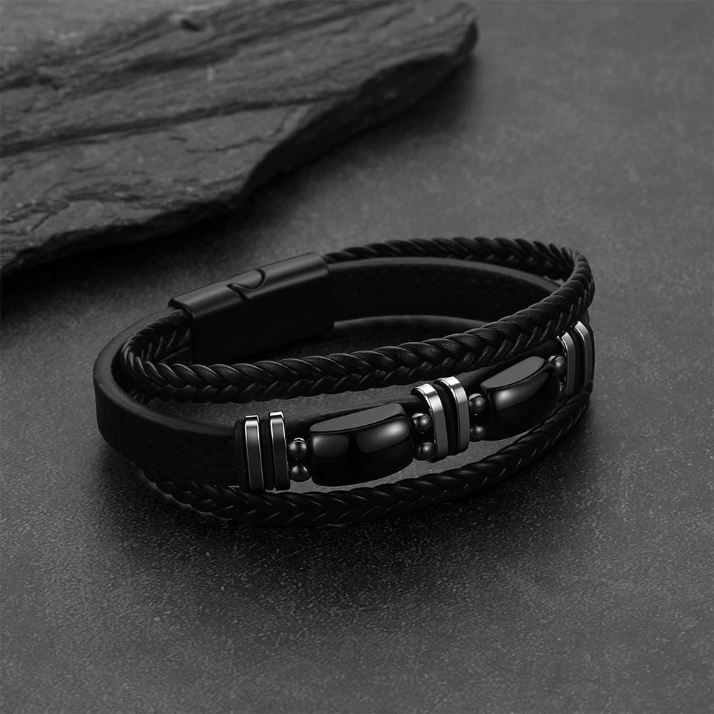 A black Obsidian Multilayer leather and braided cord bracelet with polished metallic silver-toned accents, displayed on a dark textured surface with a slate prop.