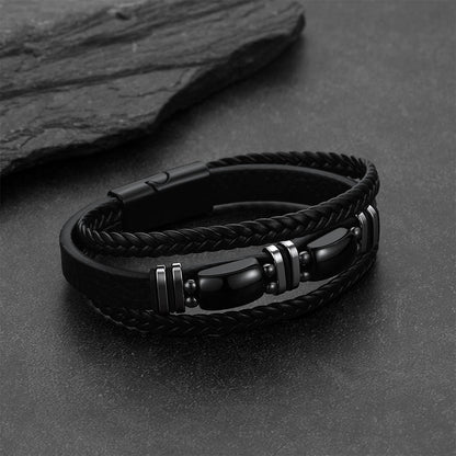 A black Obsidian Multilayer leather and braided cord bracelet with polished metallic silver-toned accents, displayed on a dark textured surface with a slate prop.