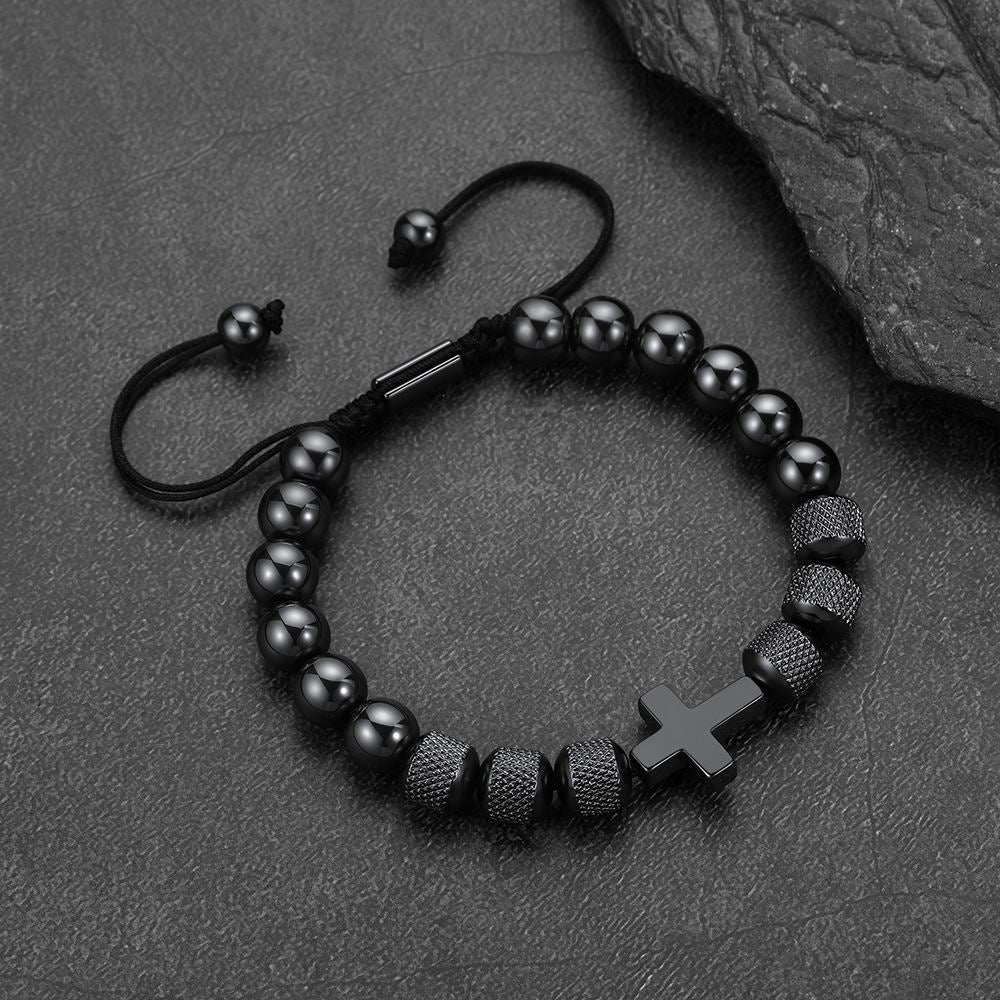 A black beaded bracelet with a matte black cross charm, textured spacer beads, and an adjustable sliding cord clasp, displayed on a dark textured slate surface.