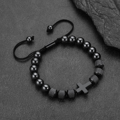 A black beaded bracelet with a matte black cross charm, textured spacer beads, and an adjustable sliding cord clasp, displayed on a dark textured slate surface.