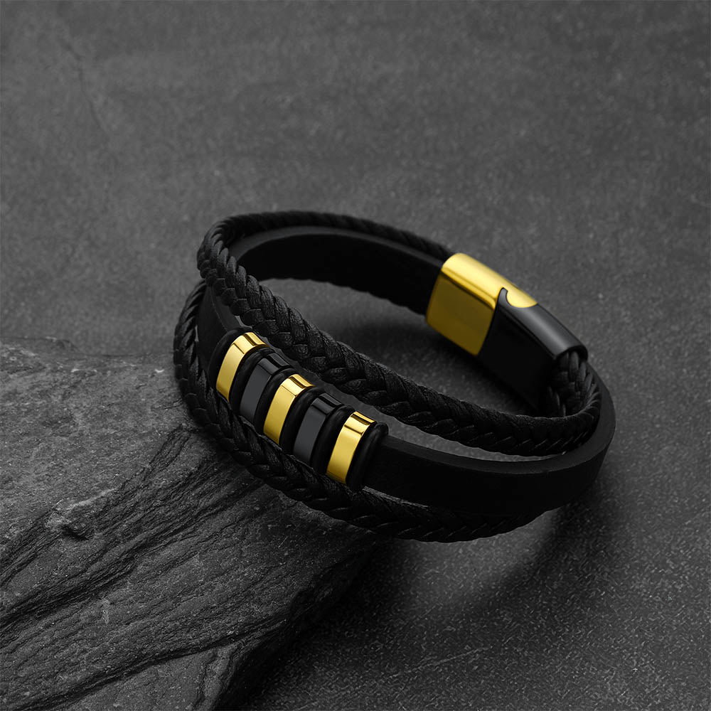 A black braided and smooth leather bracelet with polished gold-tone metal accents, displayed on a dark textured slate surface.
