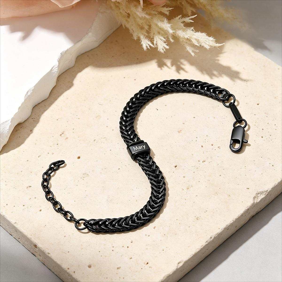 A black braided chain bracelet with a lobster clasp and a small rectangular charm engraved with 'MARTY', displayed on a beige stone slab with dried grass and soft shadows.