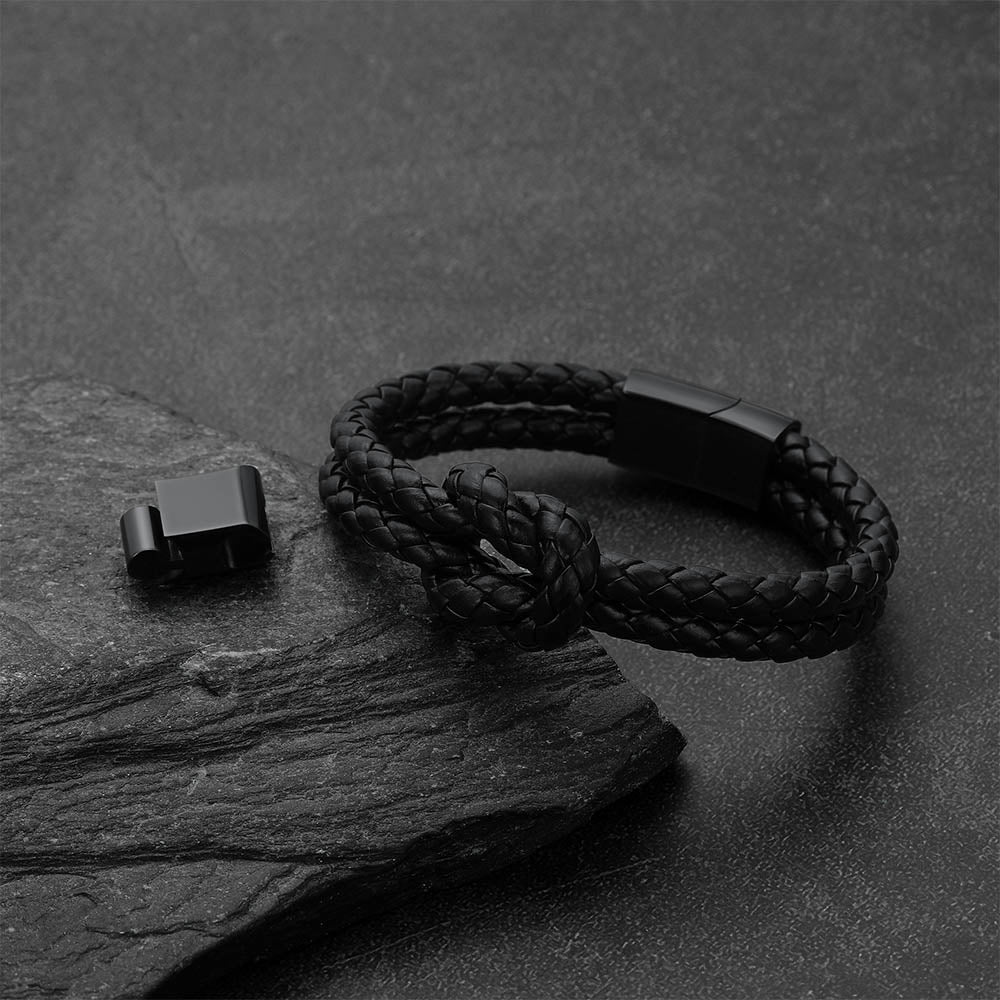 A black braided cord bracelet with a matte black metal clasp, displayed on a dark textured slate surface next to its clasp component.
