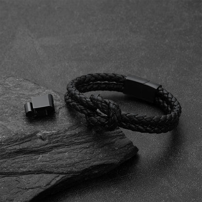 A black braided cord bracelet with a matte black metal clasp, displayed on a dark textured slate surface next to its clasp component.