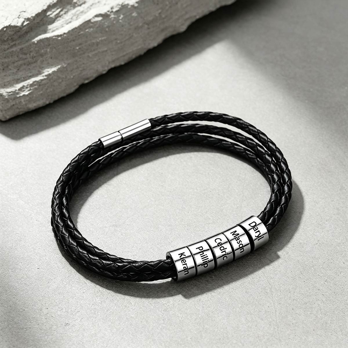 A black braided cord bracelet with four silver cylindrical name rings engraved with personalized names, displayed on a textured gray surface with a stone prop in the corner.