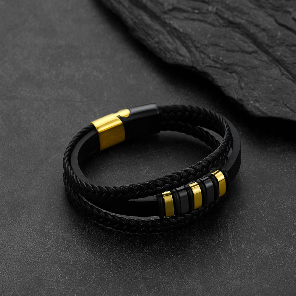 A black braided double-strand bracelet with polished gold-tone metal accents, displayed on a dark textured slate surface.