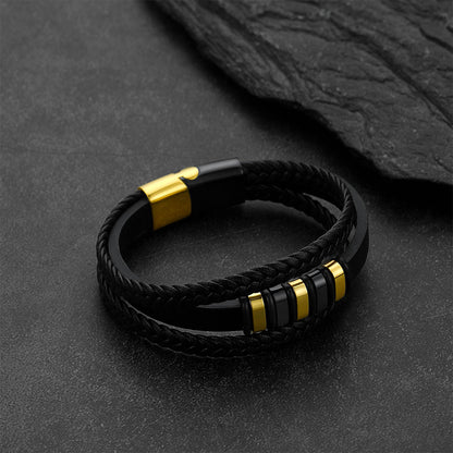 A black braided double-strand bracelet with polished gold-tone metal accents, displayed on a dark textured slate surface.