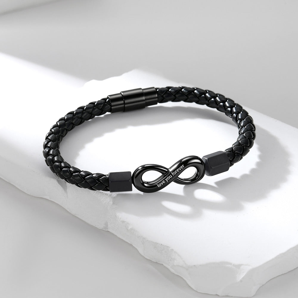 A black braided infinity leather bracelet with a matte black infinity symbol clasp engraved with 'I LOVE YOU', displayed on a white textured stone slab.