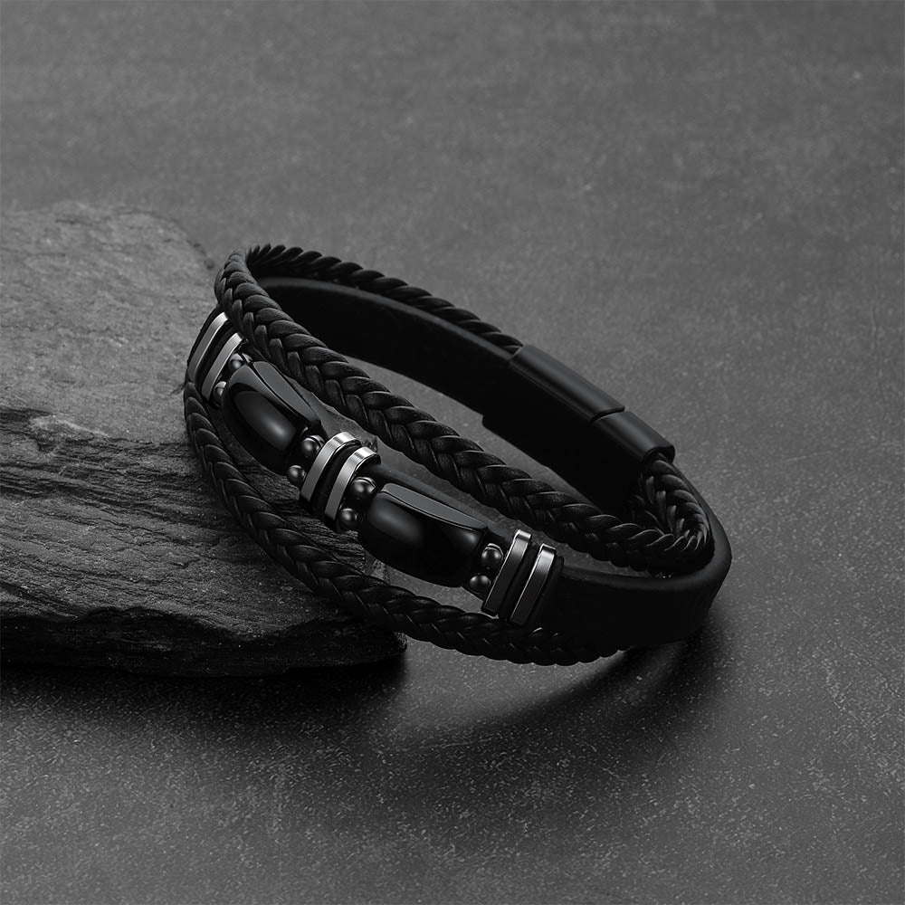 A black braided leather bracelet, displayed on a dark textured surface next to a gray stone.