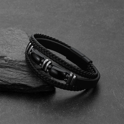 A black braided leather bracelet, displayed on a dark textured surface next to a gray stone.