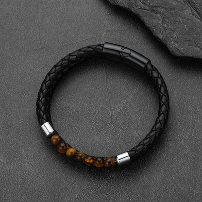 A black braided leather bracelet with a black magnetic clasp and a row of polished brown tiger's eye beads, displayed on a dark textured slate surface.