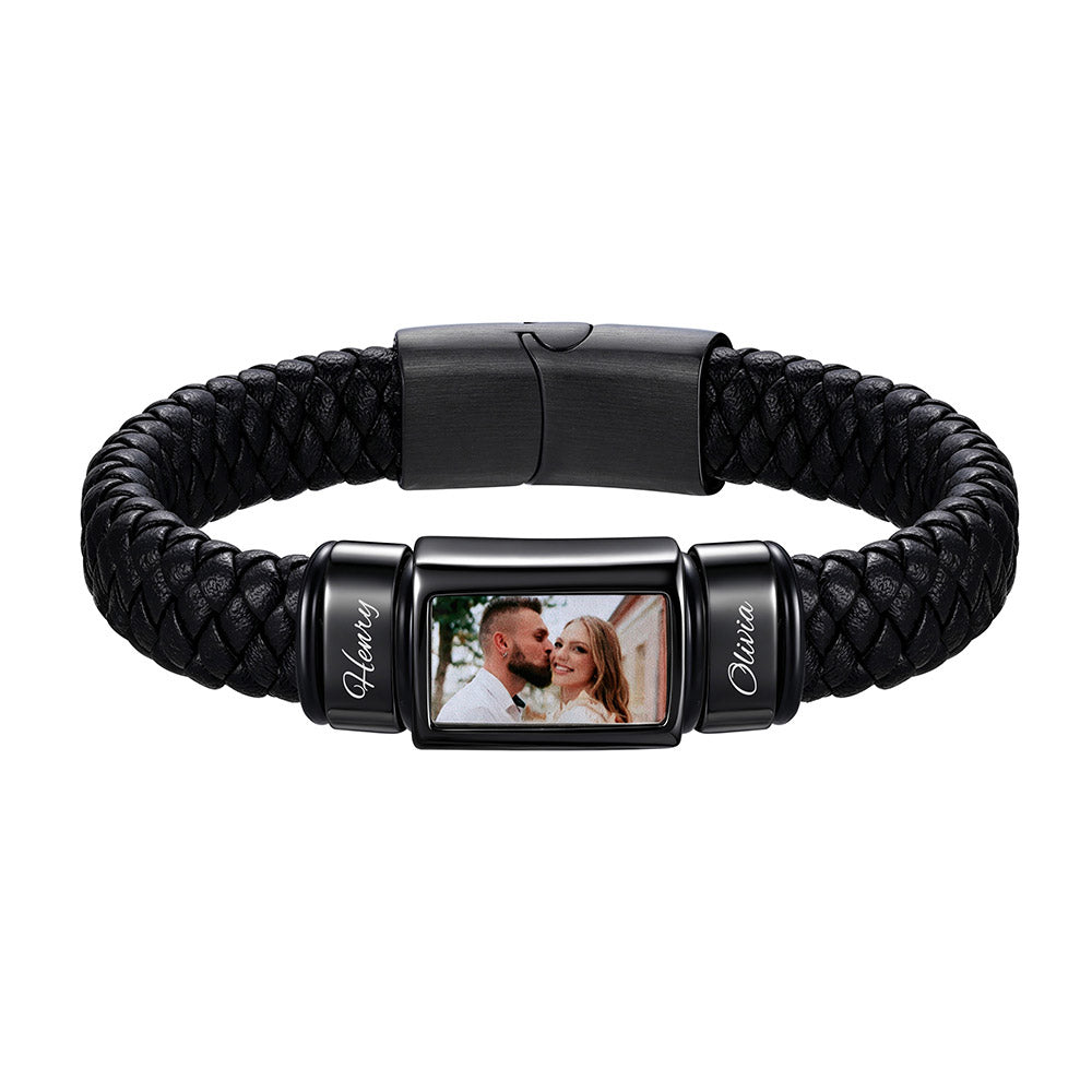 A black braided leather bracelet with a custom photo display center. The center features a rectangular black metal frame showing a custom couple's photo, flanked by two cylindrical black end caps with the names 'Homer' and 'Claudia' in white script.