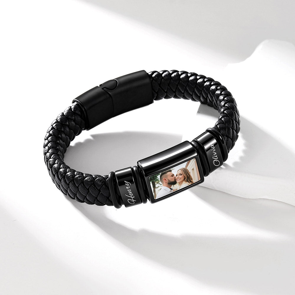 A black braided leather bracelet with a custom photo display clasp, shown on a white surface with soft shadows. The photo insert on the clasp displays a couple.