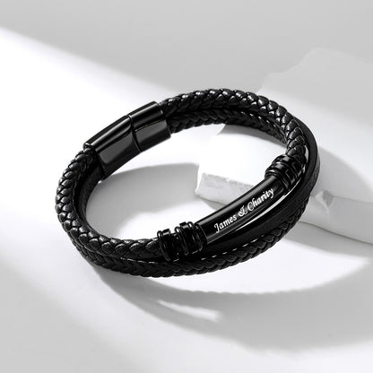 A black braided leather bracelet with a polished black metal accent bar engraved with 'James & Charity', displayed on a clean white surface with soft shadows.