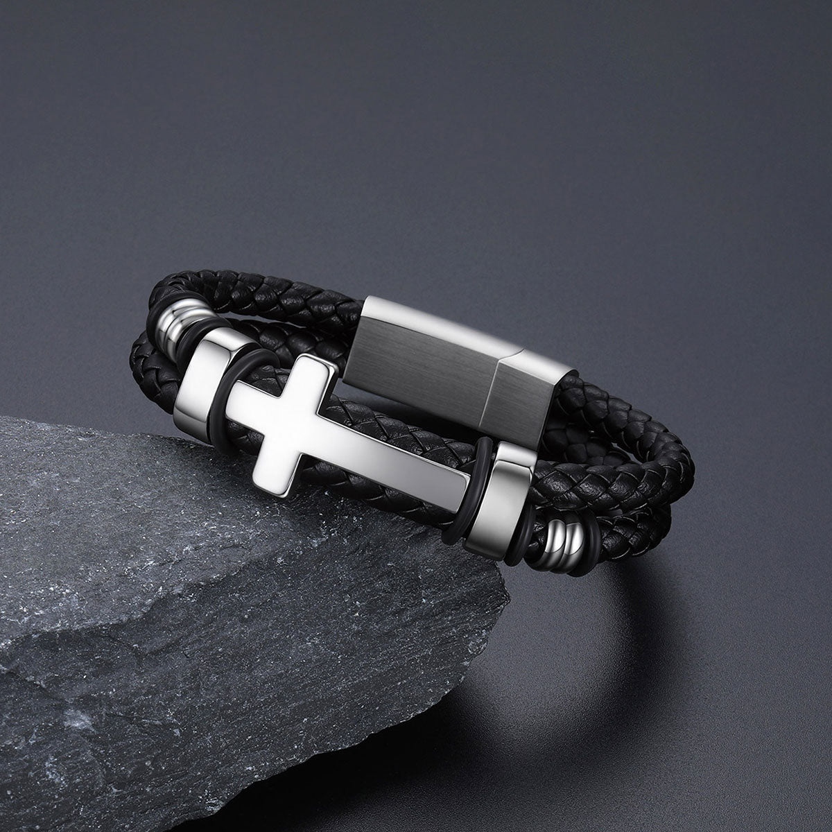 A black braided leather bracelet with a polished silver-tone cross charm and matching metal accents, displayed on a dark textured stone against a dark gray background.