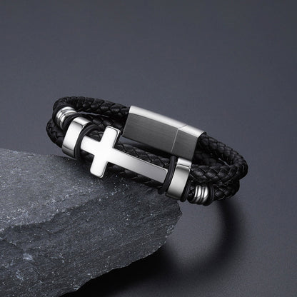 A black braided leather bracelet with a polished silver-tone cross charm and matching metal accents, displayed on a dark textured stone against a dark gray background.