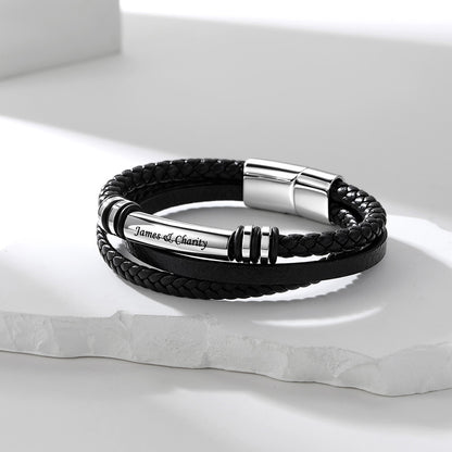 A black braided leather bracelet with a polished silver metal tube engraved with the name 'James L. Chantry', displayed on a white stone slab.