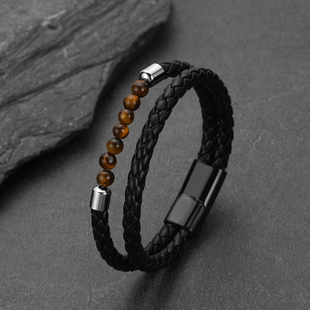 A black braided leather bracelet with a row of brown tiger's eye beads and silver-tone metal accents, displayed on a dark slate surface.