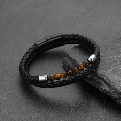 A black braided leather bracelet with a row of brown tiger's eye beads and silver-toned metal accents, displayed on a dark textured surface.