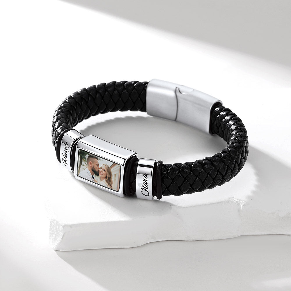 A black braided leather bracelet with a silver-toned rectangular photo plaque and a matching clasp, displayed on a white surface with soft shadows.
