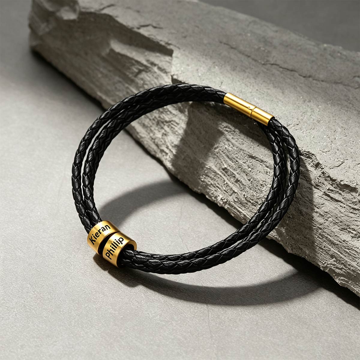 A black braided leather bracelet with gold-plated metal accents, including a custom-engraved nameplate that reads 'Keran Phillip' and a gold magnetic clasp, displayed on a textured gray stone slab.