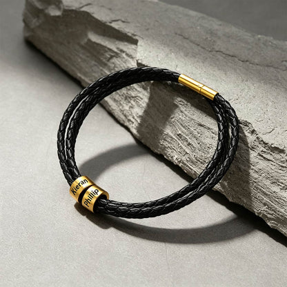 A black braided leather bracelet with gold-plated metal accents, including a custom-engraved nameplate that reads 'Keran Phillip' and a gold magnetic clasp, displayed on a textured gray stone slab.