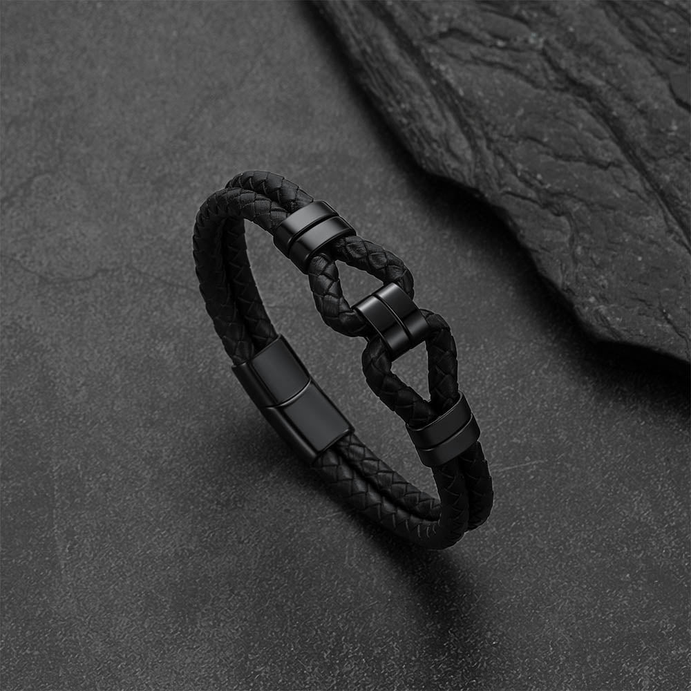A black braided leather bracelet with matte black metal accents and a knot-style clasp, photographed on a dark textured surface with a slate prop.