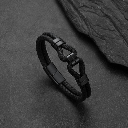 A black braided leather bracelet with matte black metal accents and a knot-style clasp, photographed on a dark textured surface with a slate prop.