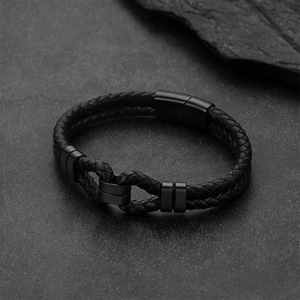 A black braided leather bracelet with matte black metal accents, displayed on a dark textured slate surface.