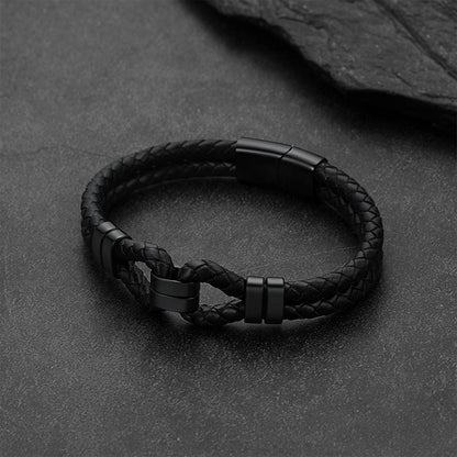 A black braided leather bracelet with matte black metal accents, displayed on a dark textured slate surface.