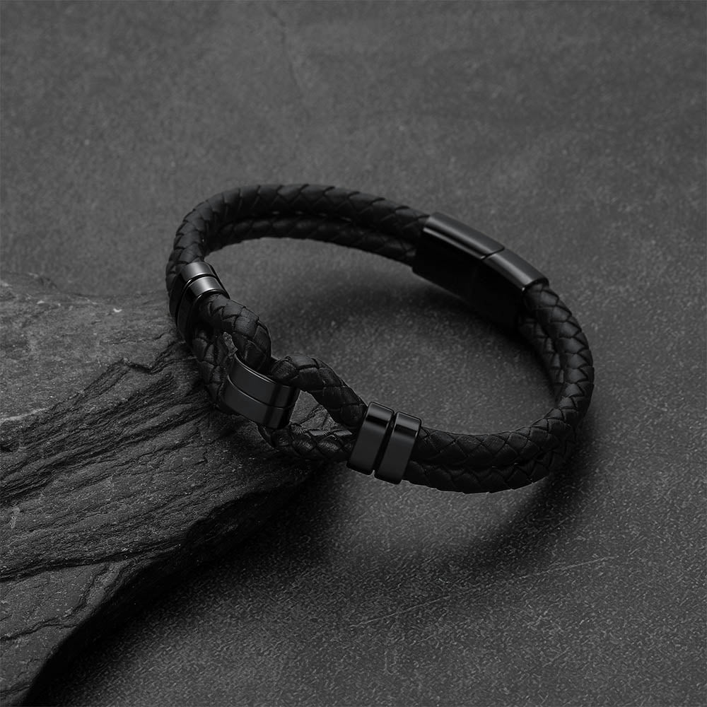 A black braided leather bracelet with matte black metal accents, resting on a dark textured stone surface.
