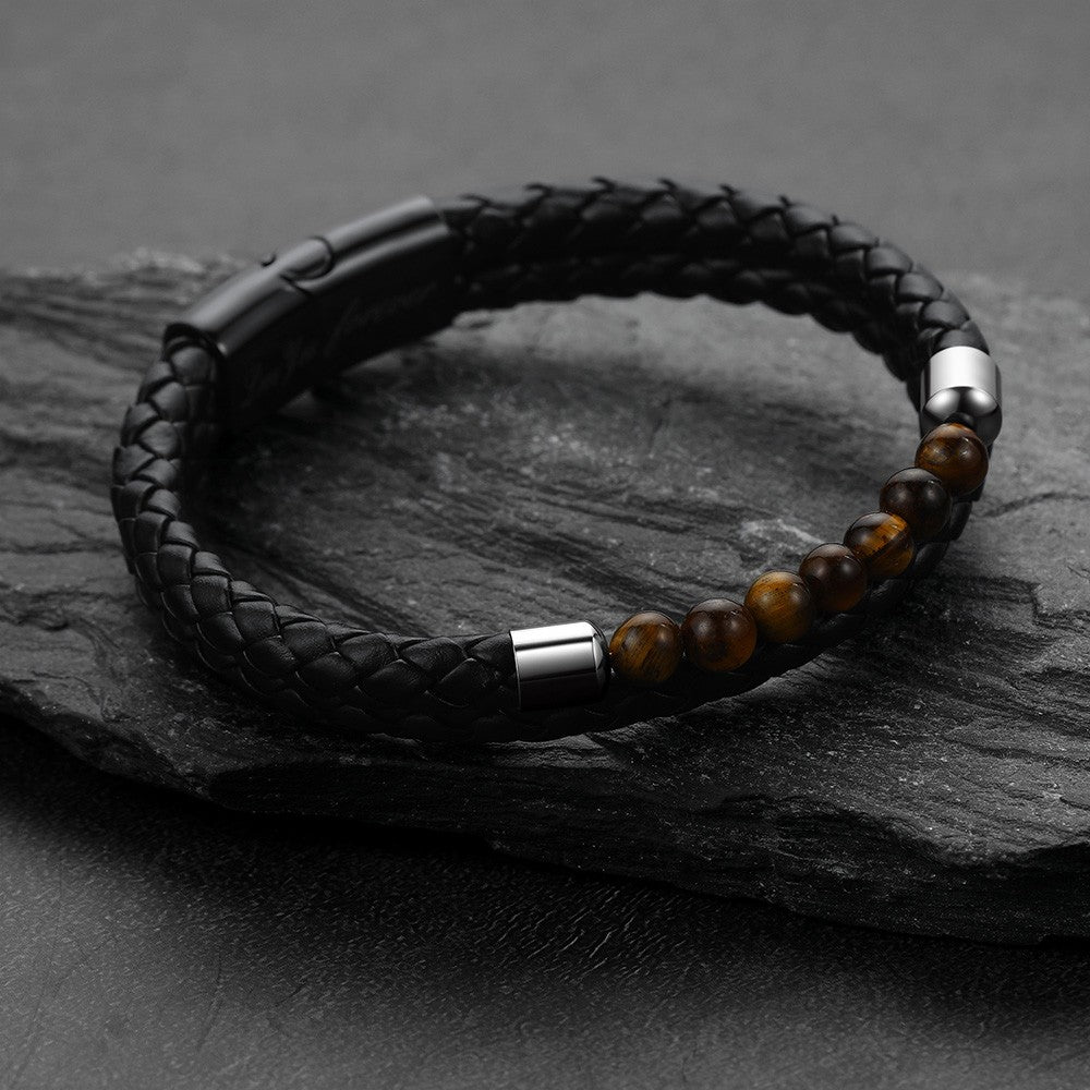 A black braided leather bracelet with polished brown tiger's eye beads and silver-toned metal accents, displayed on a dark textured slate surface.