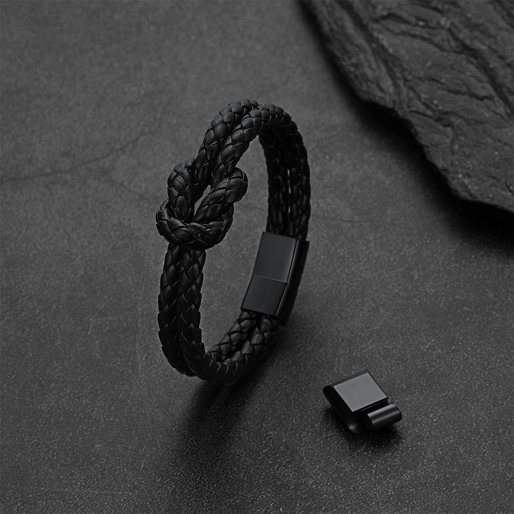 A black braided leather knot bracelet with a matte black metal clasp, displayed on a dark textured slate surface next to its clasp component.