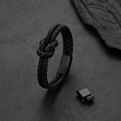 A black braided leather knot bracelet with a matte black metal clasp, displayed on a dark textured slate surface next to its clasp component.