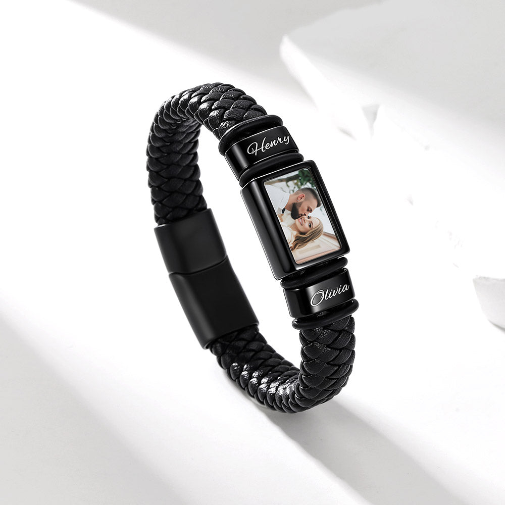 A black braided leather photo bracelet watch with a rectangular photo display showing a dog, personalized with the names 'Henry' and 'Olivia' in white script on the black metal bands, set against a clean white background with soft shadows.