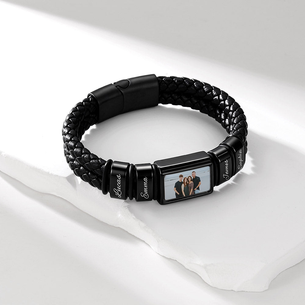 A black braided leather photo bracelet with a custom photo frame centerpiece, displayed on a white surface with soft shadows. The center of the bracelet features a rectangular photo frame showing a family portrait, flanked by three black cylindrical links engraved with the names 'Riccardo', 'Sofia', and 'Alessio'.