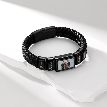 A black braided leather photo bracelet with a custom photo frame centerpiece, displayed on a white surface with soft shadows. The center of the bracelet features a rectangular photo frame showing a family portrait, flanked by three black cylindrical links engraved with the names 'Riccardo', 'Sofia', and 'Alessio'.
