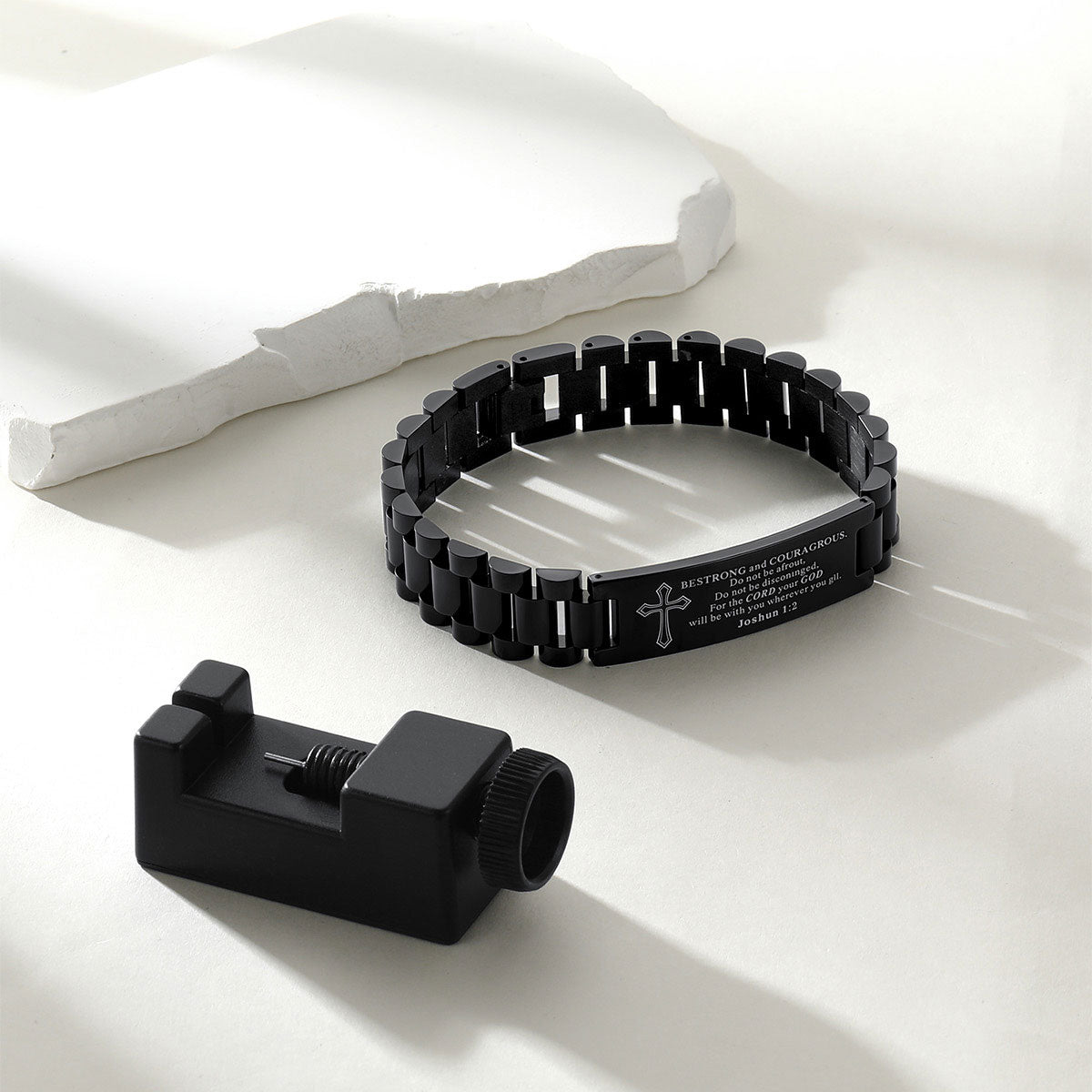 A black metal link bracelet with engraved text and a cross symbol, displayed alongside a black cylindrical tool on a clean white surface with soft shadows.