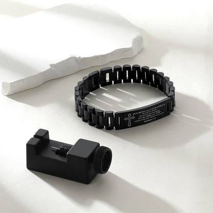 A black metal link bracelet with engraved text and a cross symbol, displayed alongside a black cylindrical tool on a clean white surface with soft shadows.