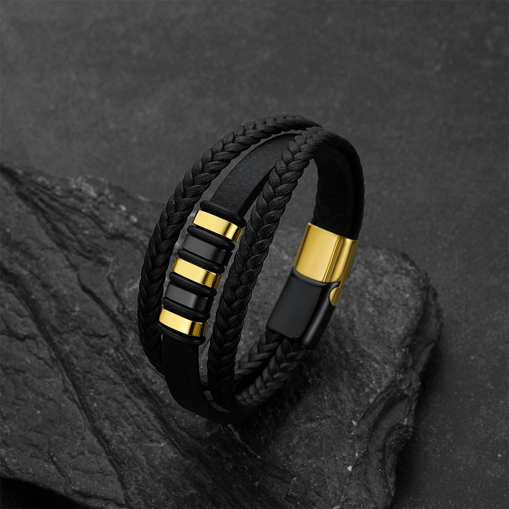 A black multi-strand bracelet with braided and smooth leather-like bands, accented by gold-tone metal details and a black magnetic clasp, displayed on a dark textured stone surface.
