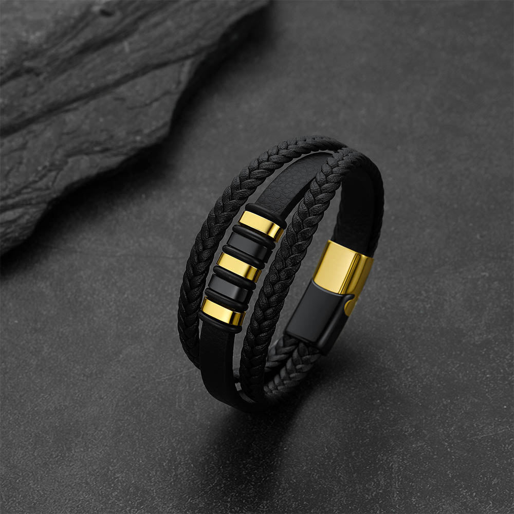 A black multi-strand leather and braided cord bracelet with polished gold-tone metal accents, displayed on a dark textured surface.