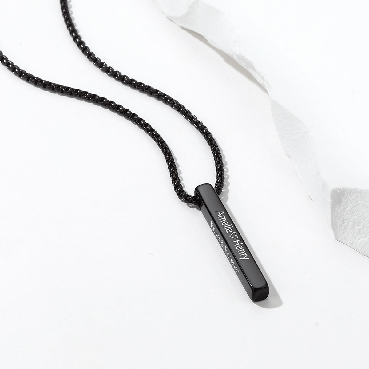 A blackengraved bar necklace with a slim black metal bar pendant engraved with 'Armen of History' in white lettering, displayed on a clean white background with a piece of crumpled paper.