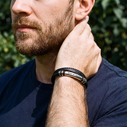 A close-up lifestyle shot of a bearded man wearing a black leather and silver-toned metal bracelet on his wrist, with a blurred green outdoor background.