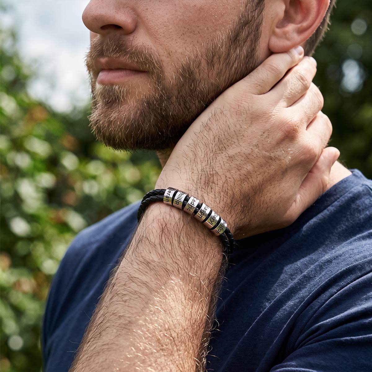 A close-up lifestyle shot of a man wearing a black cord bracelet with silver-toned metal beads engraved with 'I LOVE YOU' and 'IN LOVE' on a blurred green outdoor background.