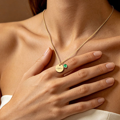 A close-up lifestyle shot of a model wearing a delicate gold chain necklace with a round gold disc pendant engraved with 'Love' and a small green gemstone charm.