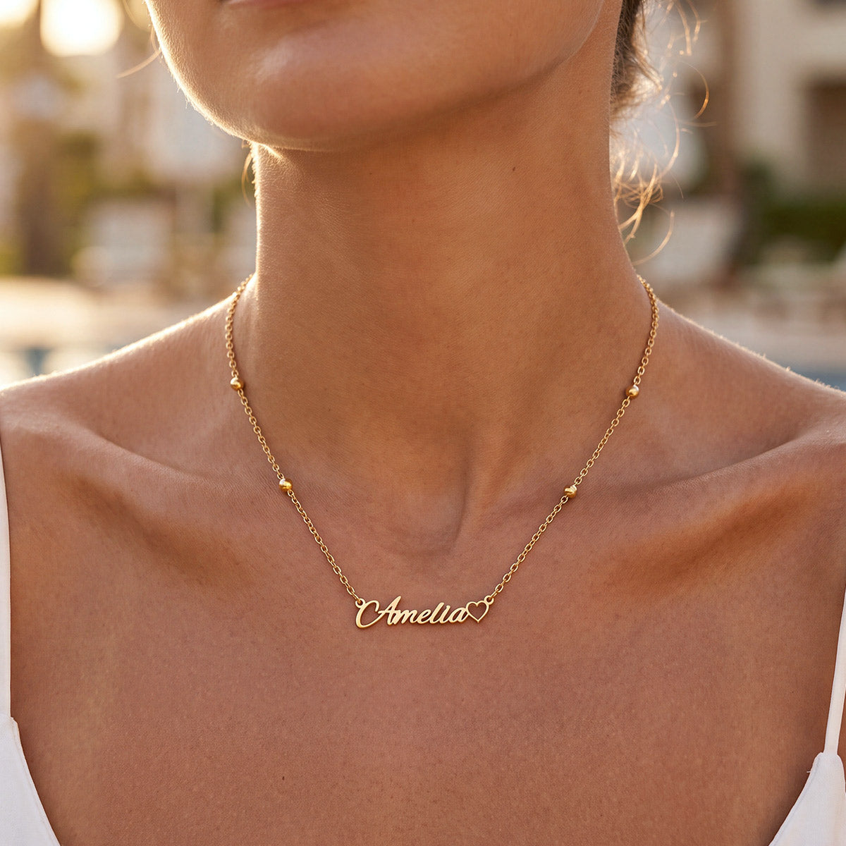 A close-up lifestyle shot of a model wearing a delicate gold name necklace with a personalized script nameplate reading 'Amelia'.
