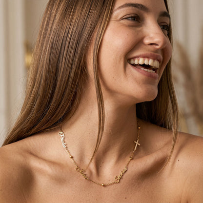 A close-up lifestyle shot of a smiling woman with long brown hair wearing a delicate gold chain necklace with custom script lettering and a small cross pendant.