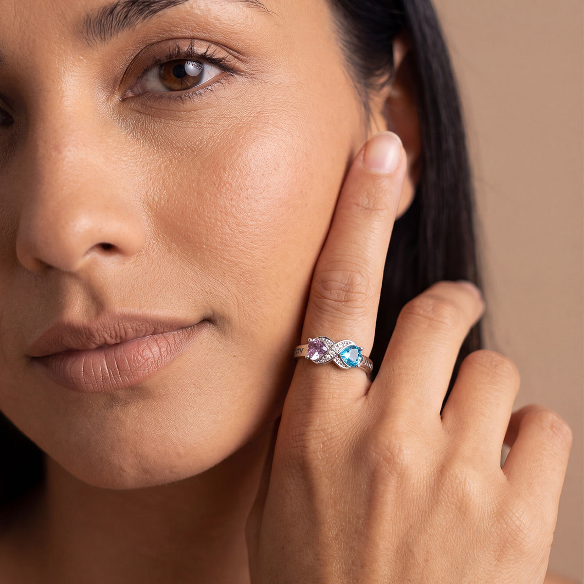 A close-up lifestyle shot of a woman wearing a delicate silver infinity ring with three small round gemstones in pink, blue, and purple, resting her hand on her cheek against a solid beige background