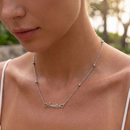 A close-up lifestyle shot of a woman wearing a delicate silver name necklace with a personalized nameplate reading 'Annelie' in cursive script, styled with small bead accents on the chain.