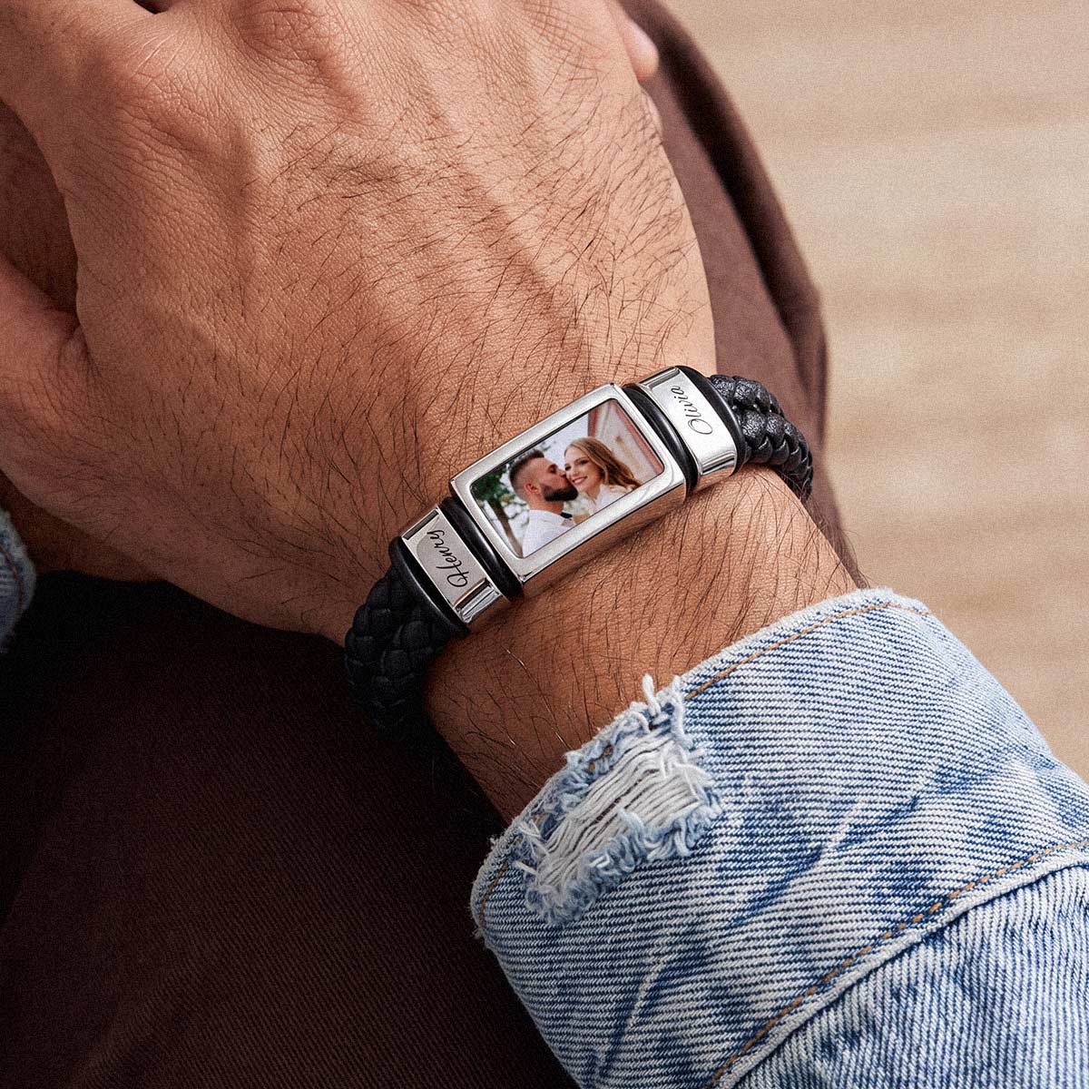 A close-up of a custom photo bracelet worn on a person's wrist. The bracelet features a black braided leather band with a polished silver-toned rectangular metal centerpiece.